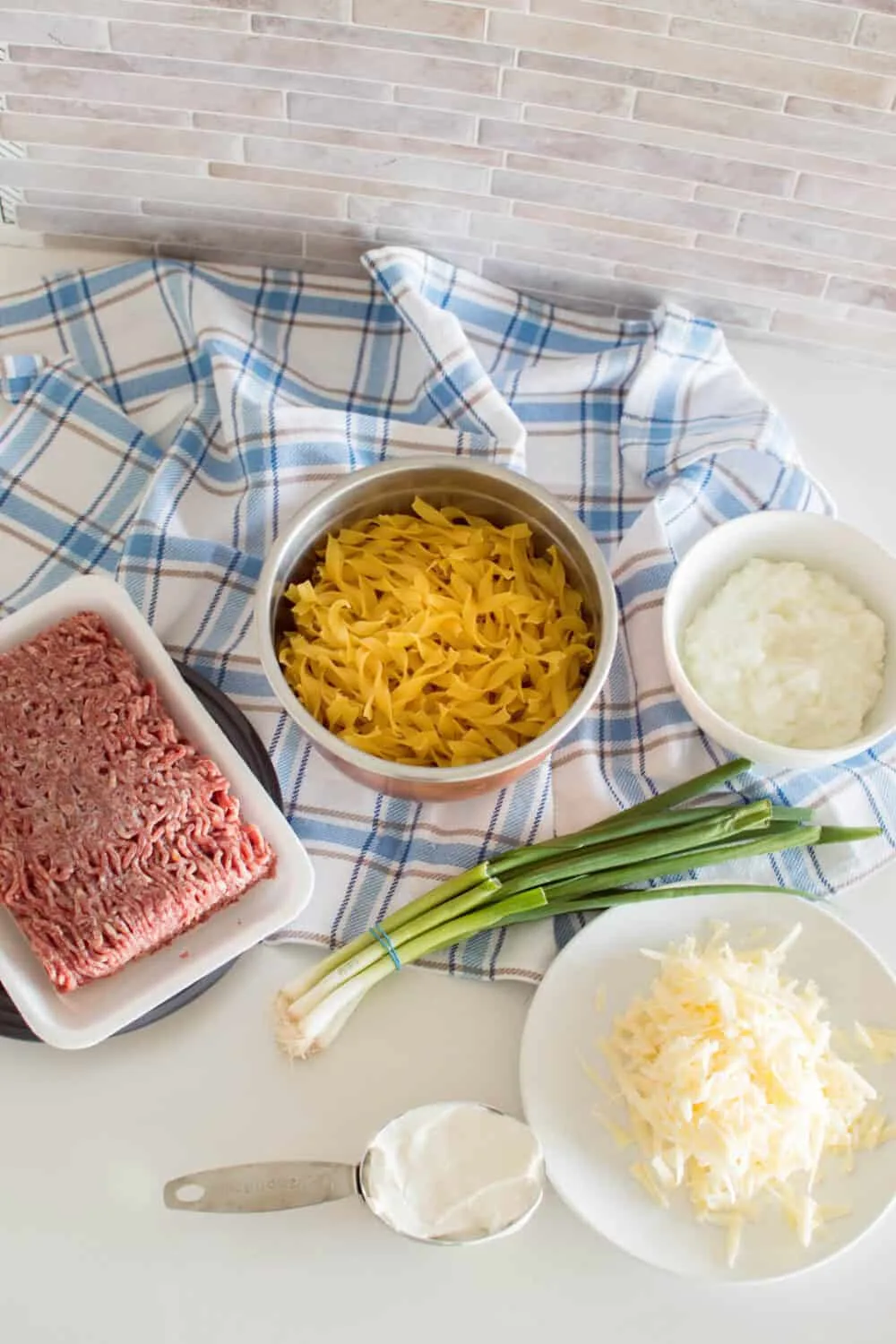 Ingredients on a white table and a blue plaid napkin to make a sour cream noodle bake