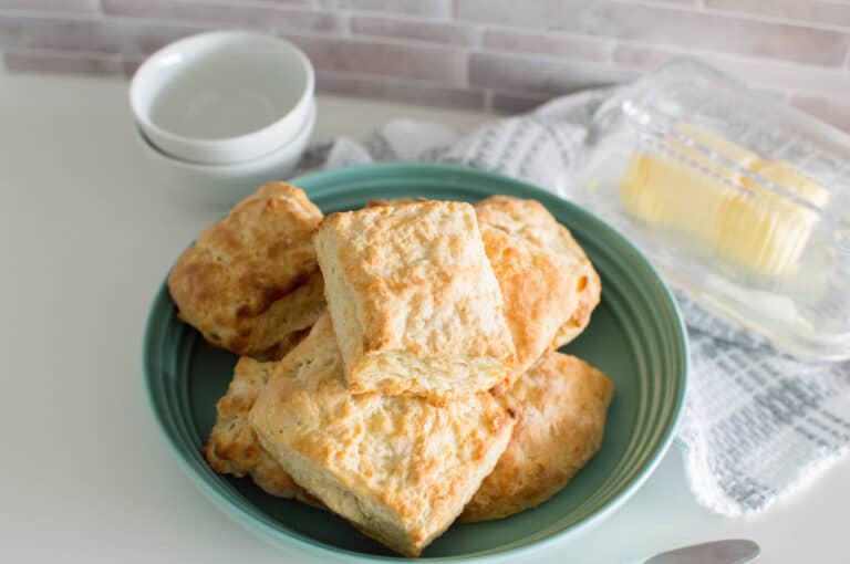 A pile of biscuits in a mint green bowl, surrounded by mini white bowls and a dish of butter