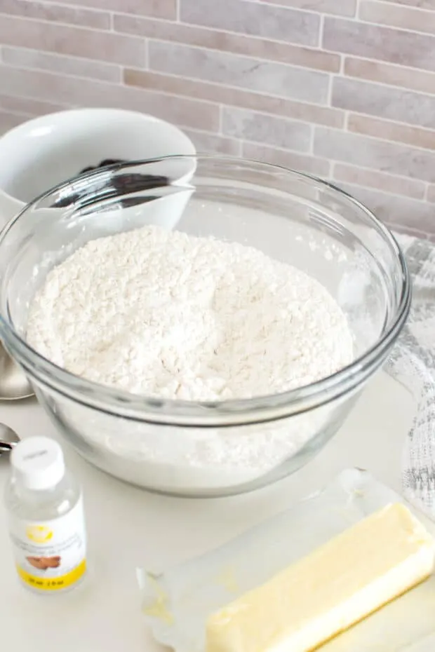 A bowl of flour in a large glass bowl surrounded by additional ingredients like butter and almond extract.