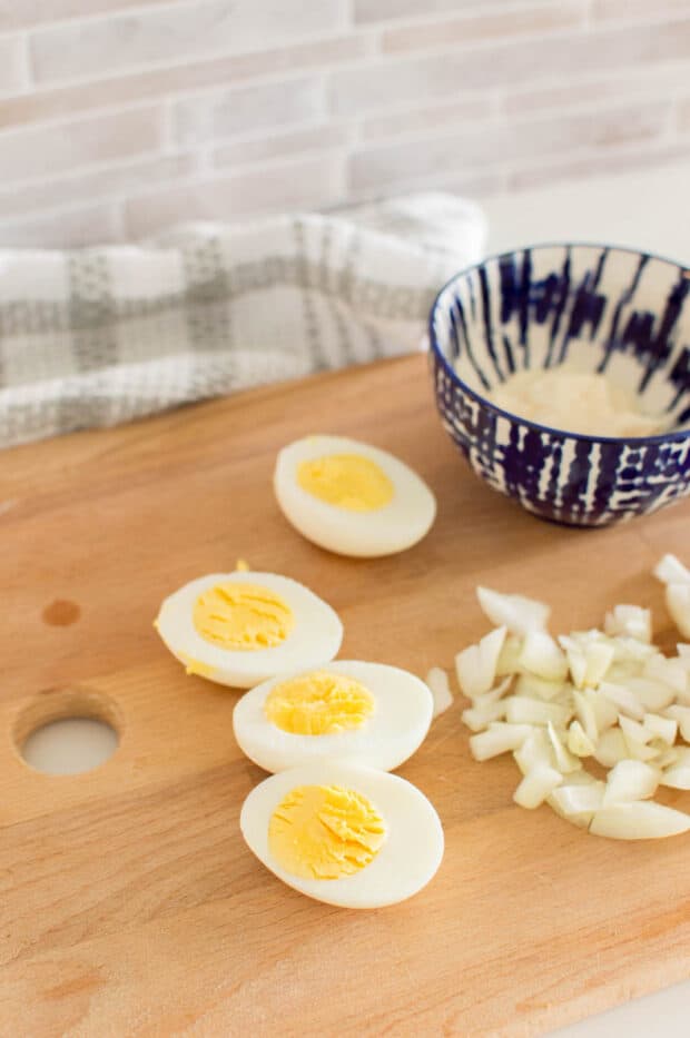 Boiled eggs cut in half next to chopped up onions and a blue bowl on a wooden cutting board