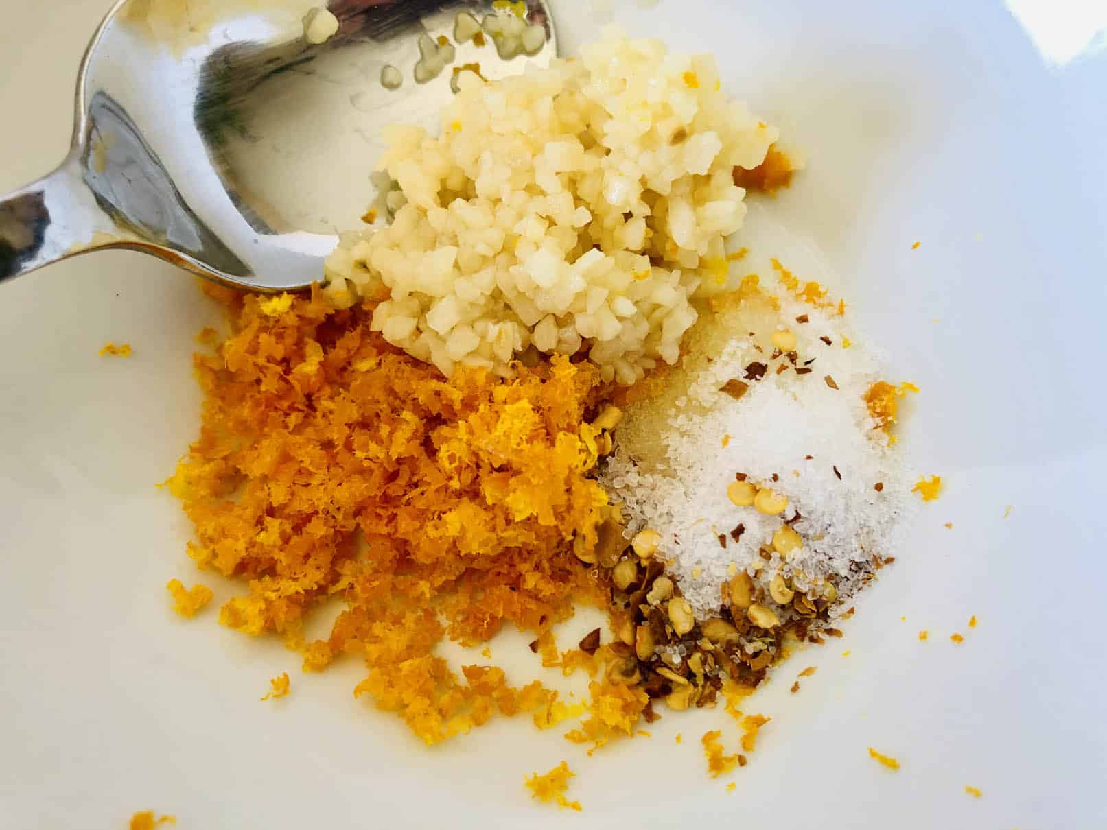Combining orange zest, minced garlic, salt and red pepper flakes in a white bowl with a spoon.