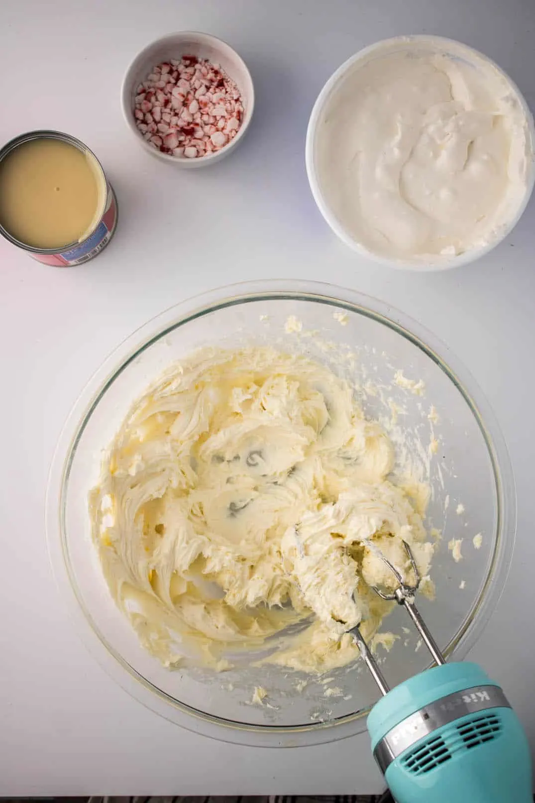 Mixing softened cream cheese and cookie sandwich fillings in a glass bowl with the help of an electric mixer. The bowl is surrounded by additional ingredients to make No-Bake Peppermint Cheesecake Jars.