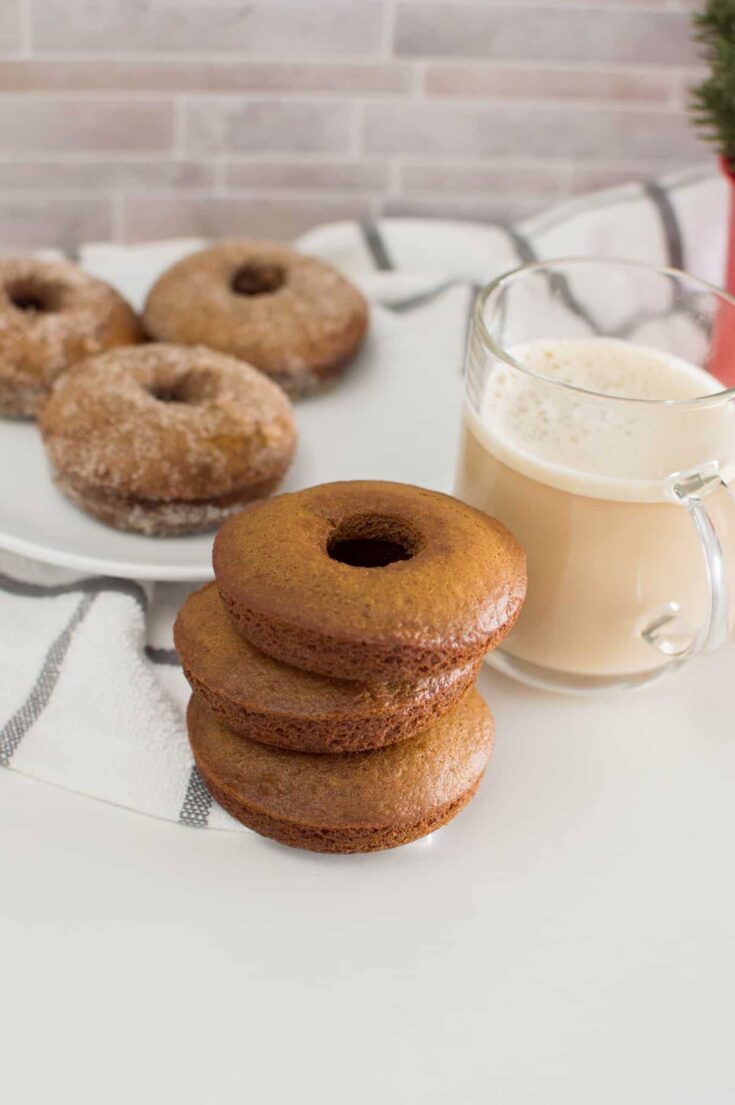 Stacked gingerbread donuts next to a cup of coffee, with more donuts in the background, sitting on a white plate
