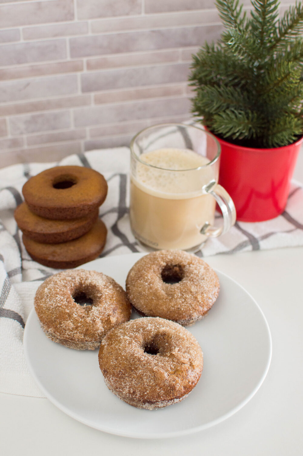 Cinnamon-sugar coated donuts on a white plate with more donuts in the background as well as a cup of coffee and a miniature Christmas tree.
