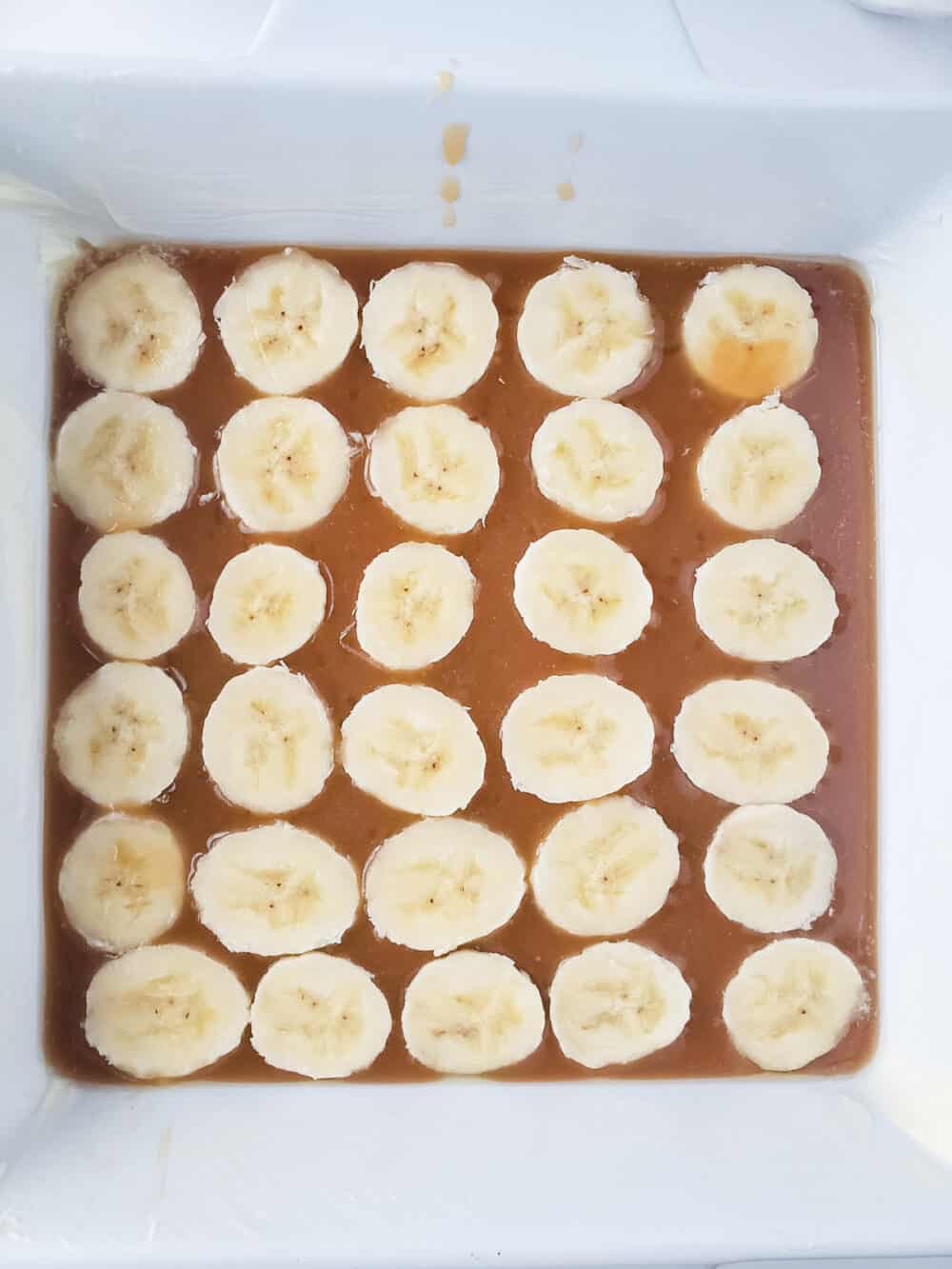 Aerial view of a homemade caramel base, topped with sliced bananas in a white casserole dish