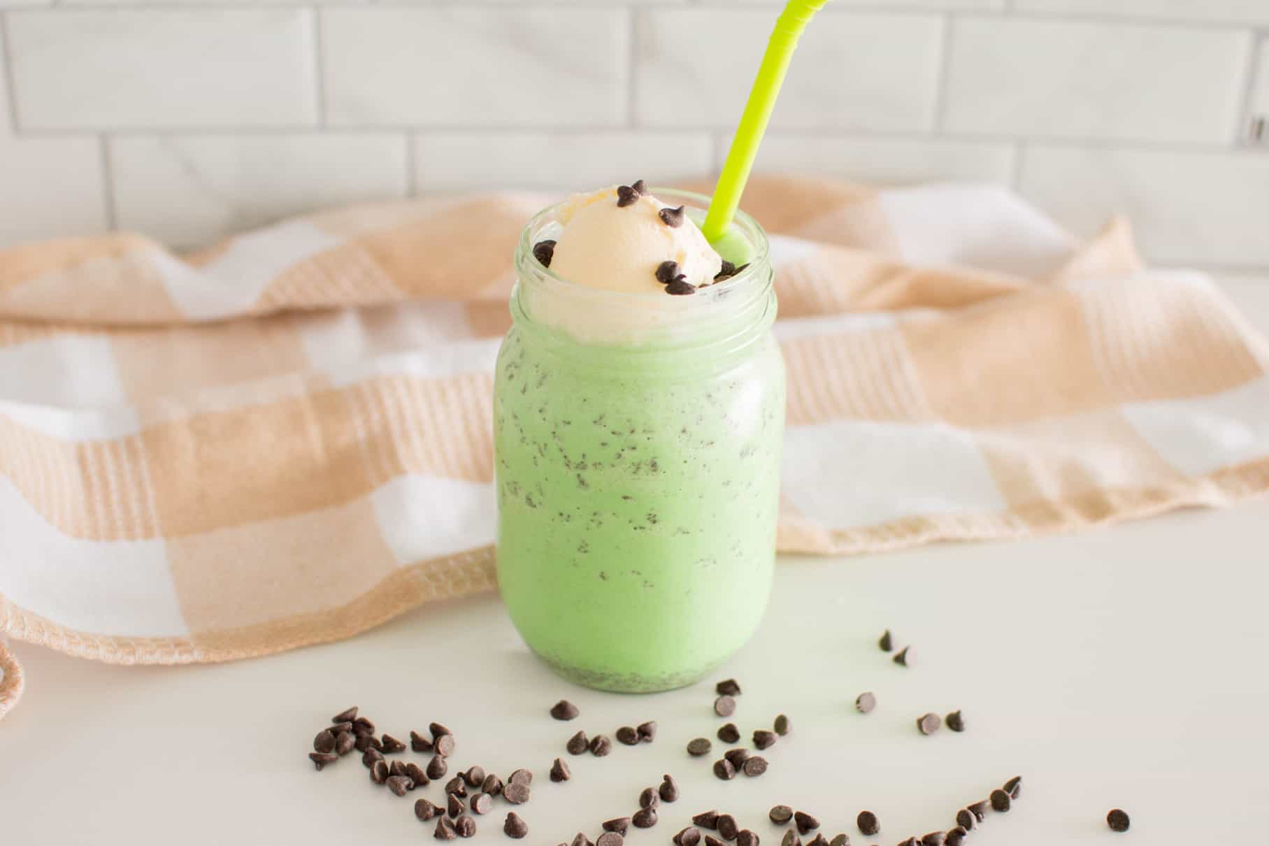 Mint Chocolate Chip Milkshake in a mason jar surrounded by chocolate chips and a plaid tea towel