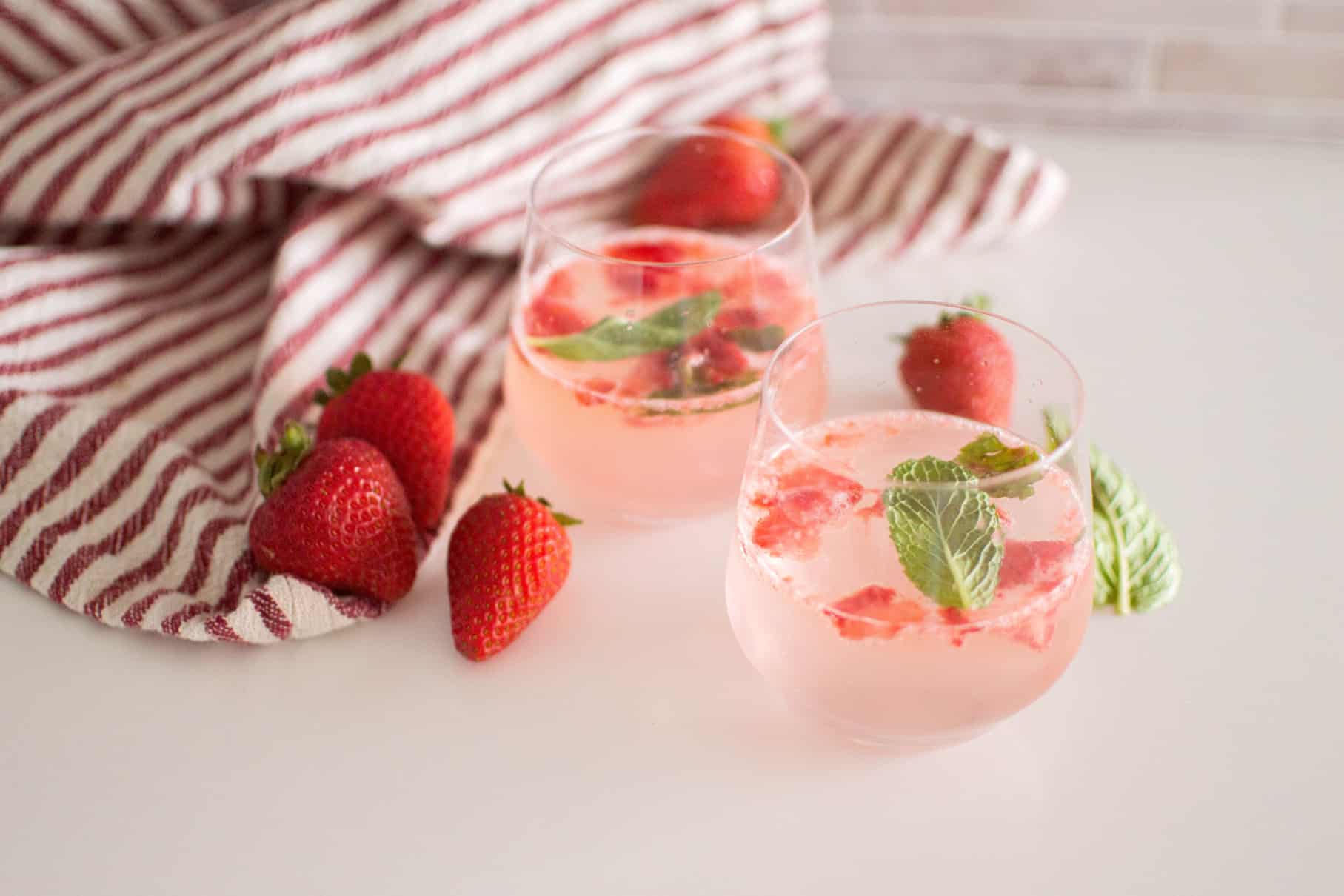 Two glasses of Mojito Mocktails with Strawberries, surrounded by fresh strawberries and a red striped towel
