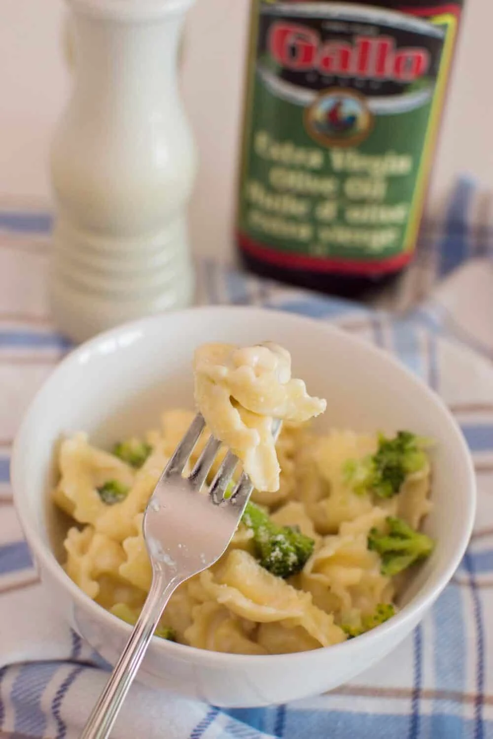 A fork with a tortellini inserted into it as the forefront of a photo of a bowl of homemade alfredo tortellini