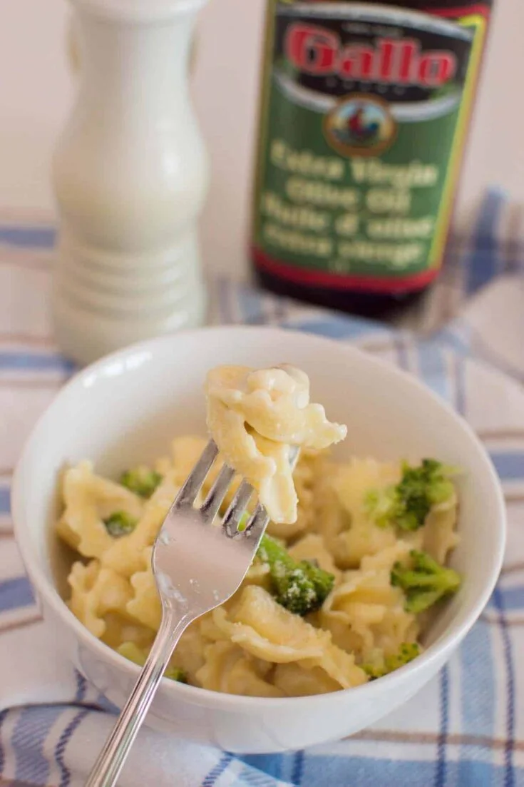 A fork with a tortellini inserted into it as the forefront of a photo of a bowl of homemade alfredo tortellini