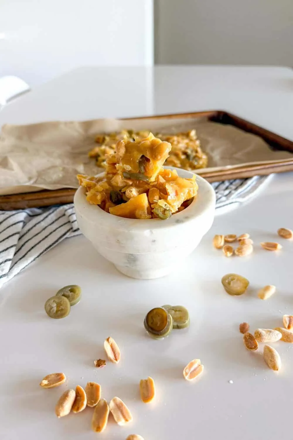 Jalapeno Peanut Brittle sitting in a marble bowl on a white table surrounded by additional pieces of peanut brittle and jalapeno