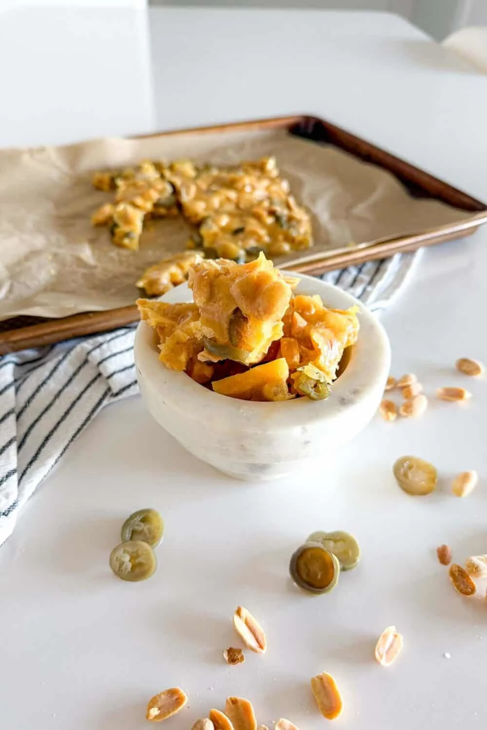 Jalapeno Peanut Brittle sitting in a marble bowl on a white table surrounded by additional pieces of peanut brittle and jalapeno