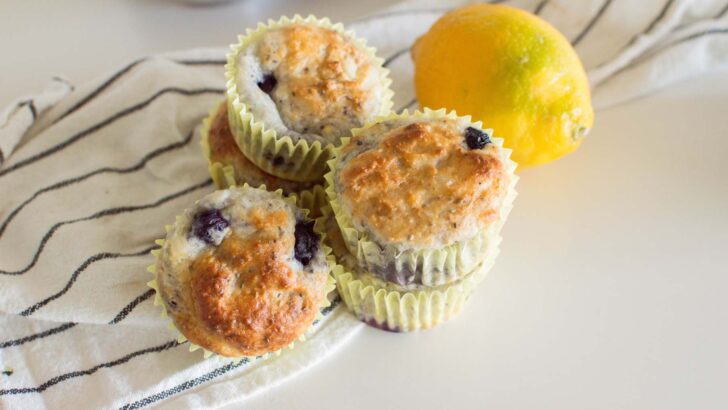 A stack of Lemon Poppyseed Blueberry Muffins on a white table surrounded by a lemon and a tea towel