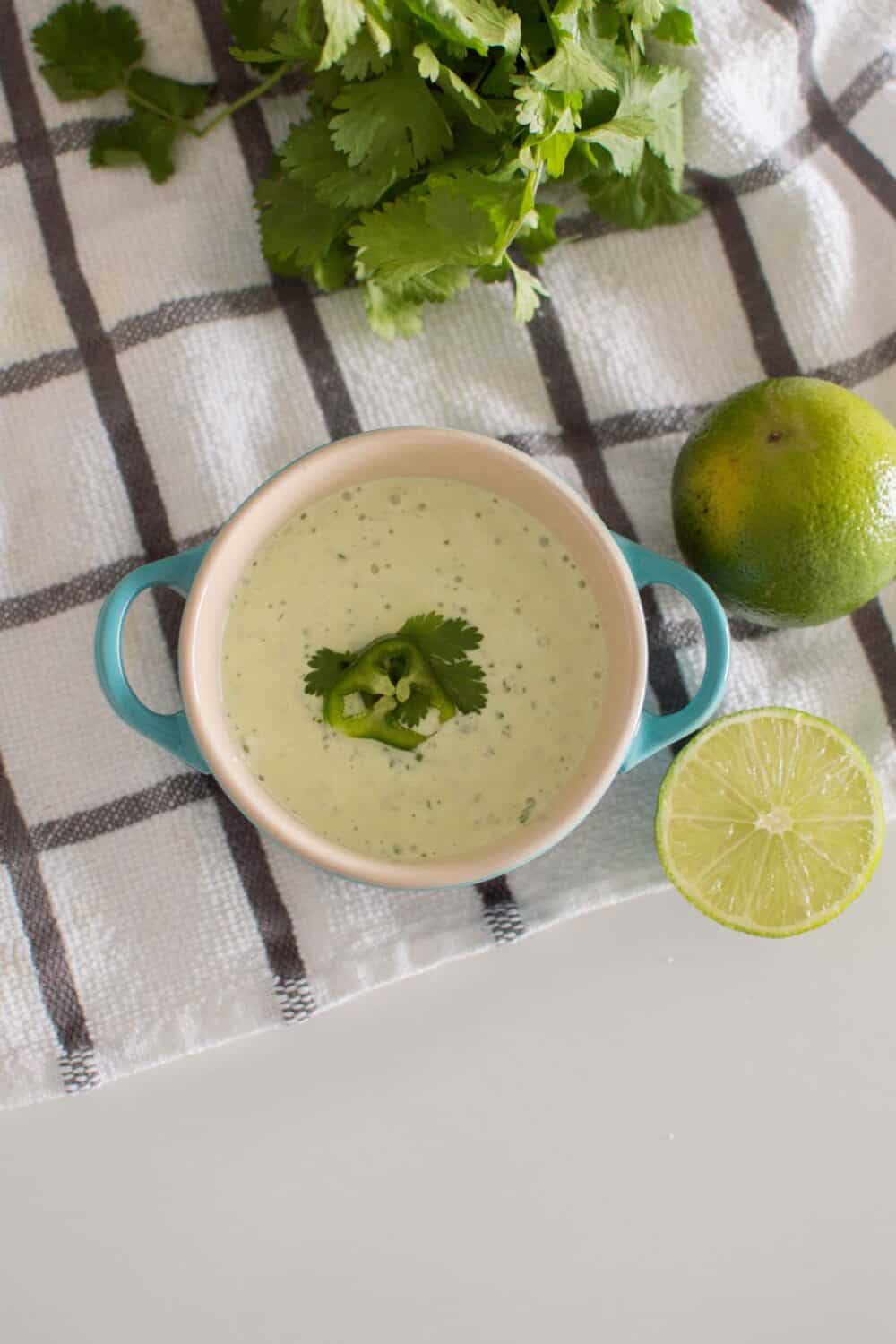 Bird's eye view of Jalapeno Aioli served in a blue serving dish, surrounded by limes and fresh cilantro